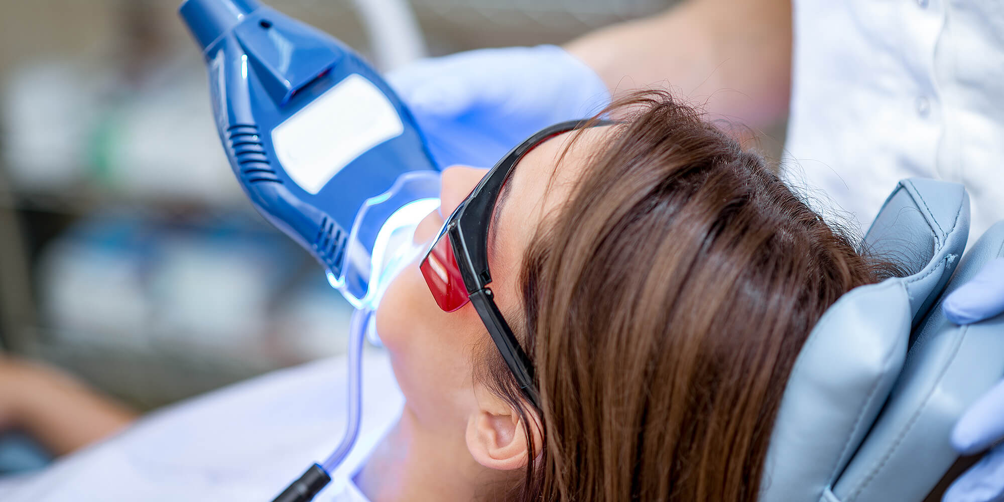 Patientin im Zahnarztstuhl mit Schutzbrille; ein blaues LED-Gerät beleuchtet den Mund während einer Zahnbehandlung bzw. Zahnaufhellung.