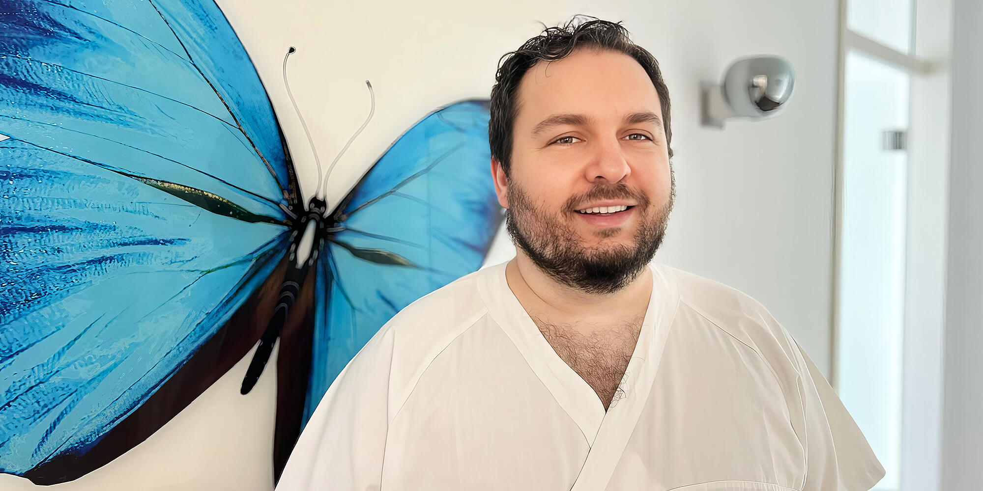 Zahnarzt in grüner Praxisbekleidung steht lächelnd in modernem Behandlungsraum mit Zahnarztstuhl und großem Fenster mit Ausblick.