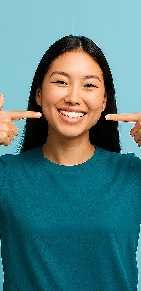 Junge Frau mit langem dunklem Haar, die mit beiden Fingern auf ihr breites, gesundes Lächeln zeigt.
