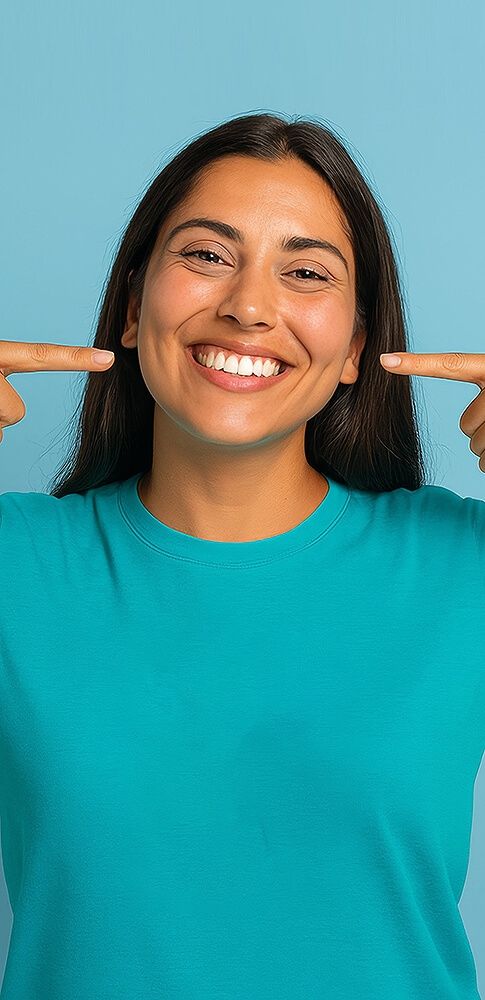 Frau mit langem dunklem Haar, die breit lächelt und mit beiden Fingern auf ihre Zähne zeigt.