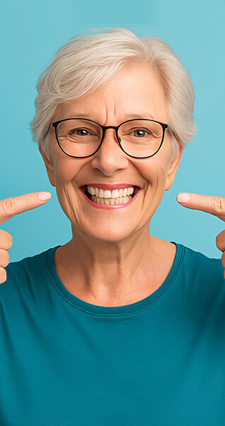 Ältere Frau mit Brille lächelt und zeigt mit beiden Zeigefingern auf ihre Zähne.