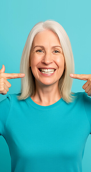 Frau mit langen grauen Haaren lächelt und zeigt mit beiden Zeigefingern auf ihre Zähne.